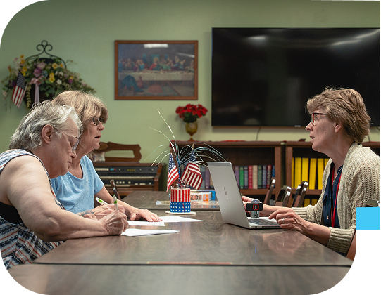 Three older women engaged in a discussion with a staff member at Senior Connections, seated around a table with a laptop and patriotic centerpiece in a cozy activity room.